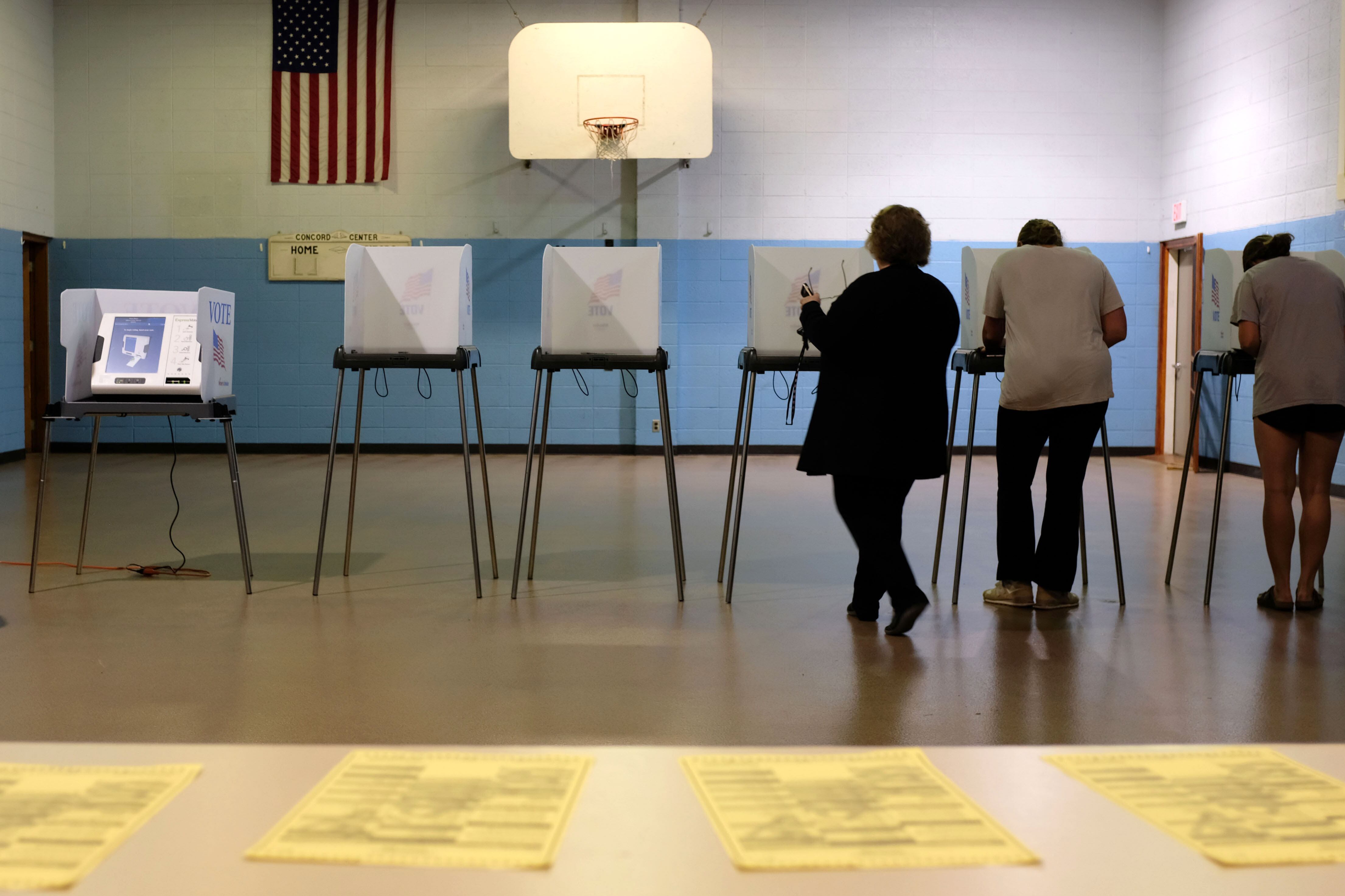 Three people stand at voting booths in a gym with a table full of yellow ballots in the foreground.