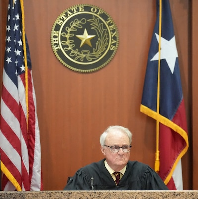 A judge sits in a courtroom between an American flag and a Texas flag with the state seal and a wood panel wall in the background.