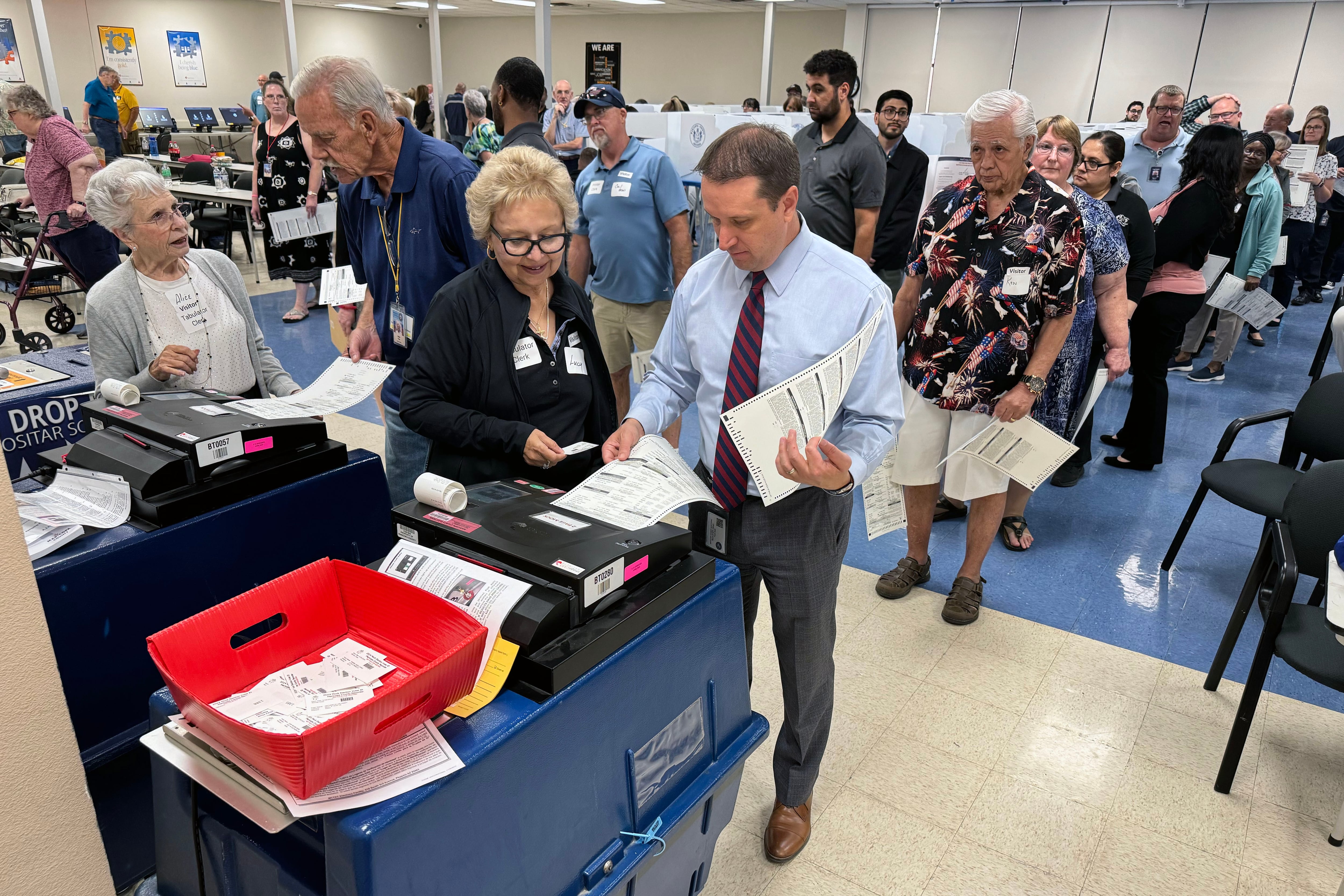 Two people work at a voting machine while a large group of people in the background in a room.