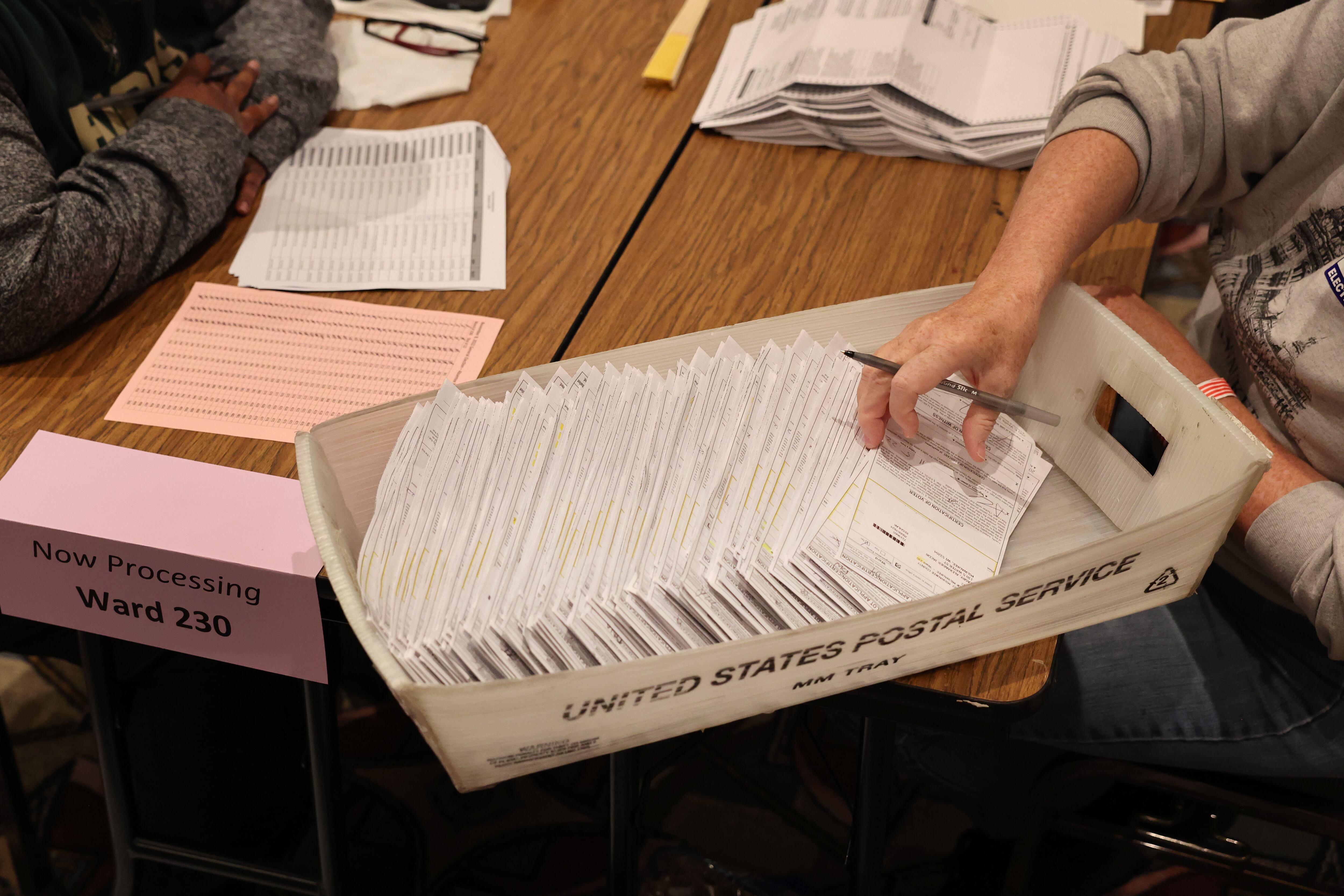 A bird's eye view of a person sitting at a wooden table with their hand in a box sorting through absentee ballots.