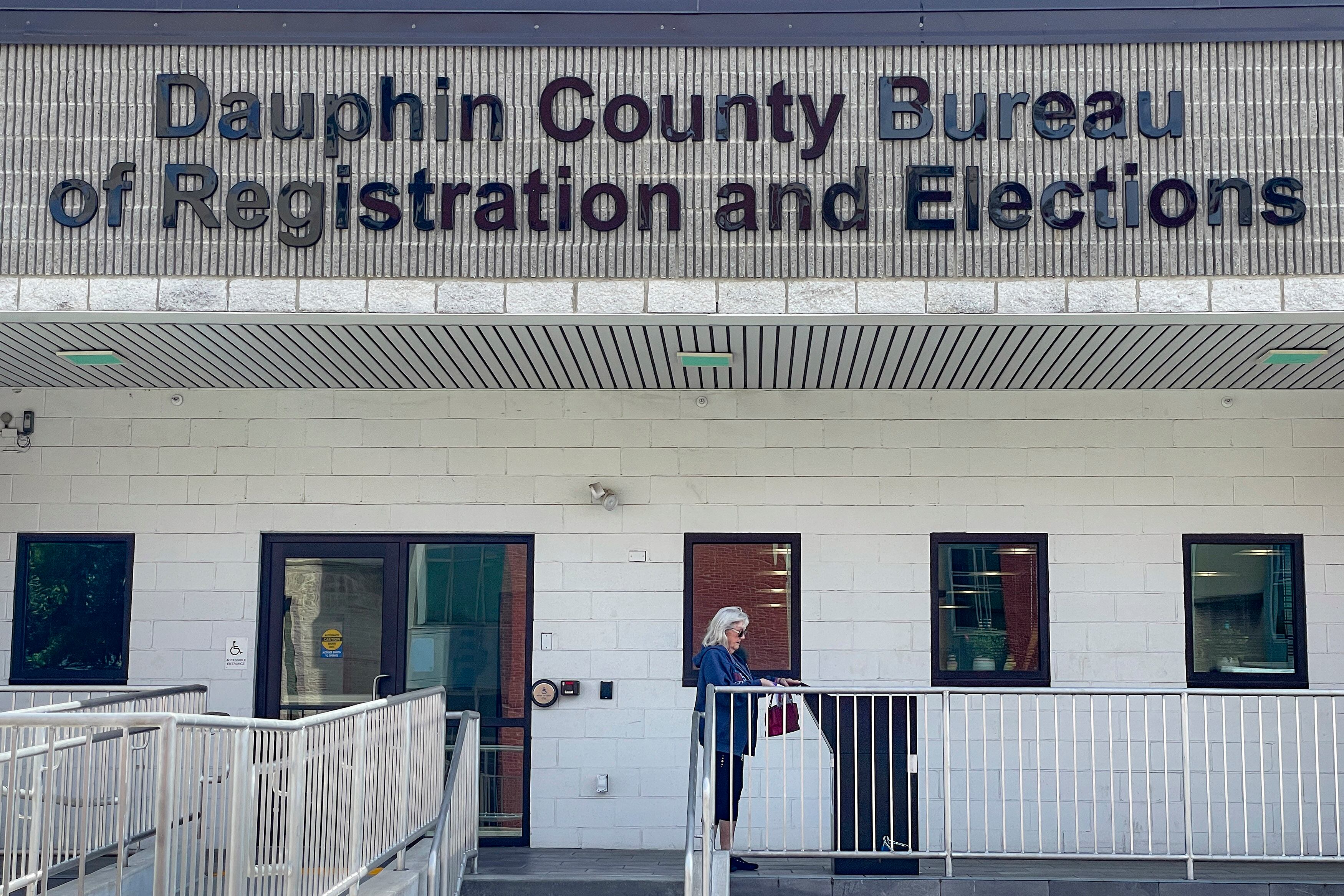 A person with short white hair stands next to a ballot drop box in front of a building with the words "Dauphin County Bureau of Registration and Elections."